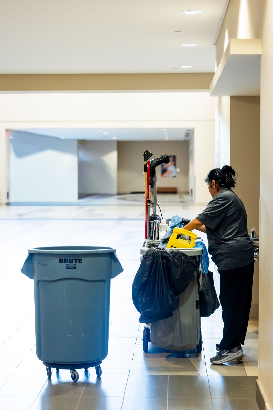 Janitorial cleaner pushing a cleaning cart down an office hallway